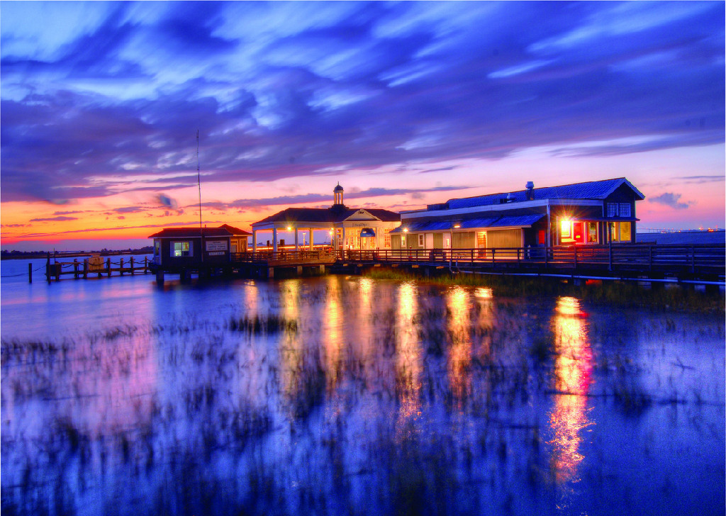 A waterfront building complex on a pier at dusk, illuminated with warm lights reflecting on still water under a dramatic sky